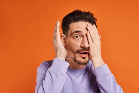 Foto de Hombre sorprendido con barba en suéter púrpura ocultar la cara con la mano sobre fondo naranja, humor juguetón - Imagen libre de derechos