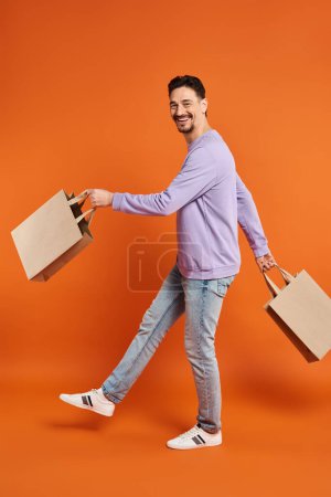 full length of happy bearded man in casual attire walking with shopping bags on orange background