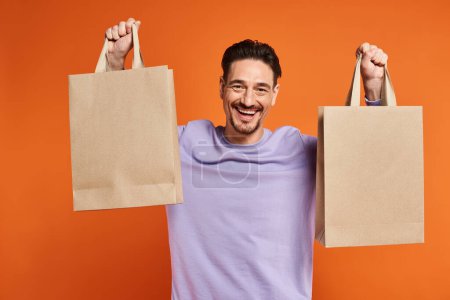 happy bearded man in casual attire holding shopping bags on orange background, consumerism