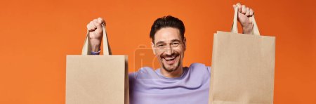 happy bearded man in casual attire holding shopping bags on orange background, consumerism banner