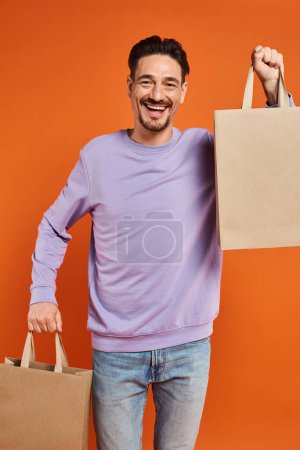 fröhlicher bärtiger Mann in lässiger Kleidung mit Einkaufstaschen auf orangefarbenem Hintergrund, Konsumdenken