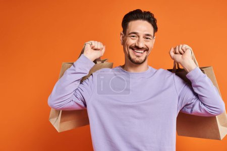 positiver bärtiger Mann in lässiger Kleidung mit Einkaufstaschen auf orangefarbenem Hintergrund, Konsumdenken