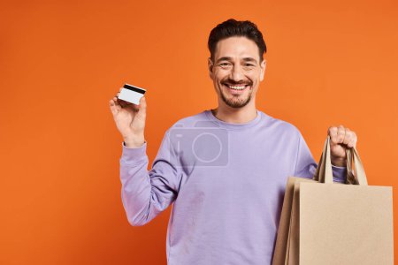 positive bearded man in casual attire holding shopping bags and credit card on orange background