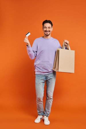 fröhlicher bärtiger Mann in lässiger Kleidung mit Einkaufstaschen und Kreditkarte auf orangefarbenem Hintergrund
