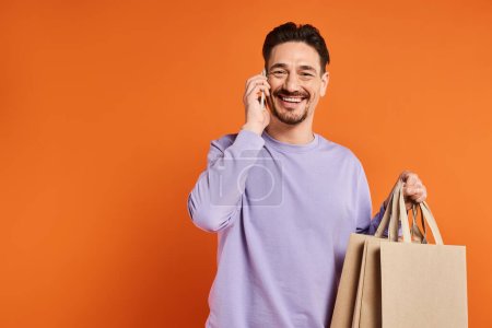 Sonriente hombre hablando por teléfono y llevando bolsas de compras sobre fondo naranja, consumidor