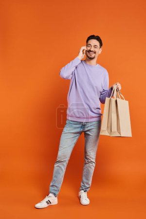 hombre alegre hablando por teléfono y llevando bolsas de compras sobre fondo naranja, consumidor