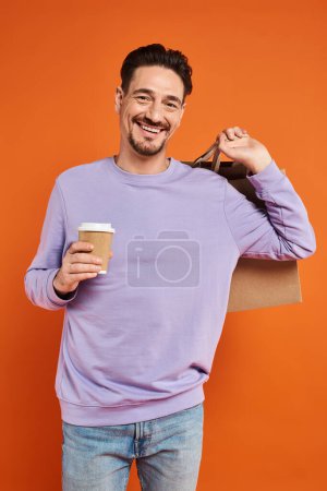 happy and bearded man holding shopping bag and coffee to go on orange background, consumerism