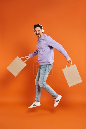 Foto de Hombre alegre en auriculares inalámbricos levitando con bolsas de compras sobre fondo naranja, compra - Imagen libre de derechos