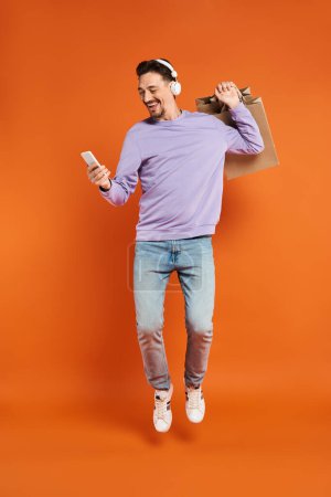 Aufgeregter Mann mit Kopfhörern schwebt mit Einkaufstaschen herum und hält Smartphone auf orangefarbenem Hintergrund