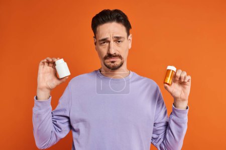 bärtiger Mann in lila Sweatshirt mit Flaschen mit Pillen auf orangefarbenem Hintergrund, Medikamente