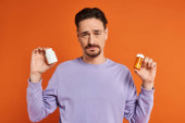 bearded man in purple sweatshirt holding bottles with pills on orange background, medication Poster #692776850
