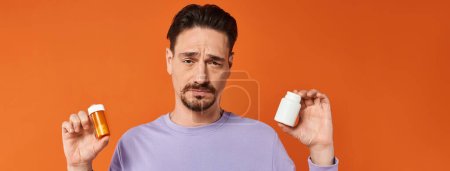 bearded man in purple sweatshirt holding bottles with pills on orange background, medication banner Mouse Pad 692776902
