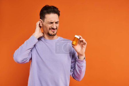 bärtiger Mann in lila Sweatshirt mit Flasche mit Pillen auf orangefarbenem Hintergrund, Medikamente