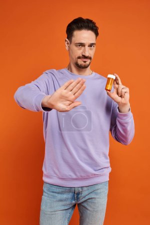 bärtiger Mann in lila Sweatshirt mit Flasche mit Pillen und Stopp auf orangefarbenem Hintergrund