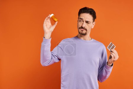 bärtiger Mann in lila Sweatshirt mit Flasche mit Pillen und Blisterverpackung auf orangefarbenem Hintergrund