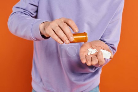 bärtiger Mann in lila Sweatshirt, der Pillen auf orangefarbenem Hintergrund in die Hand schüttet, Medikamente