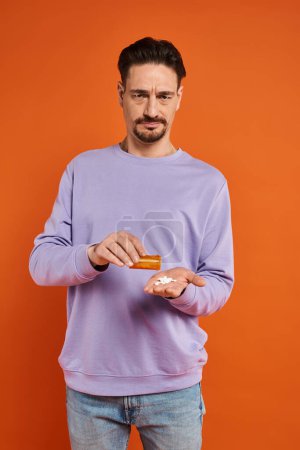 bärtiger Mann im lila Sweatshirt, der auf orangefarbenem Hintergrund Medikamente in die Hand schüttet, Pillen