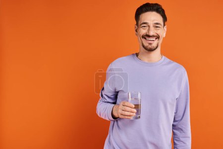 happy bearded man in purple sweatshirt holding glass of water on orange background, hydration