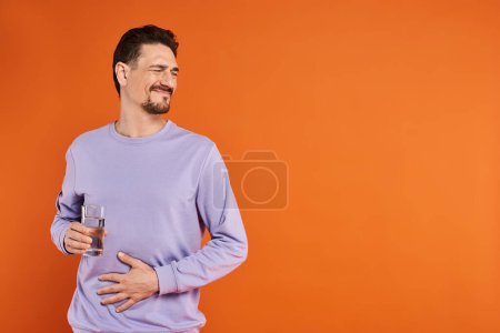 bearded man in purple sweatshirt feeling stomachache and holding glass of water on orange background