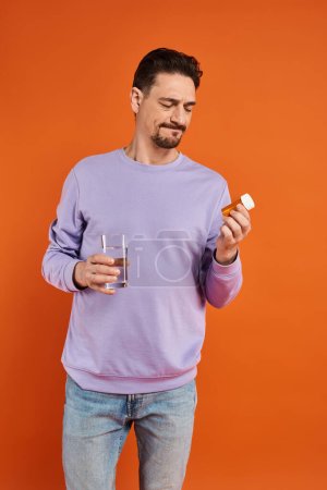 bearded man in purple sweatshirt holding glass of water and bottle with pills on orange background
