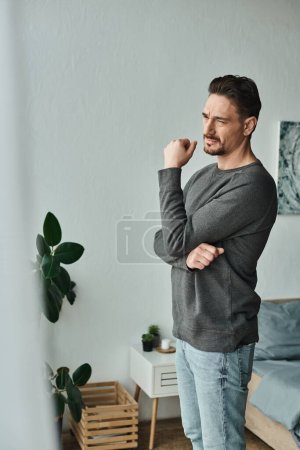 Foto de Hombre reflexivo y barbudo en jersey gris mirando hacia otro lado mientras está de pie en casa, profundamente en el pensamiento - Imagen libre de derechos
