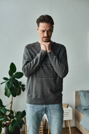 depressiver und bärtiger Mann in lässiger Heimkleidung, die Hände vor dem Gesicht auf grauem Hintergrund, traurig