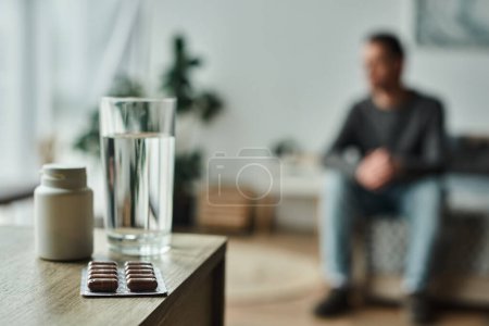 Medikamente in Flasche und Blisterverpackung neben Glas Wasser auf dem Tisch und verschwommener Mann im Hintergrund