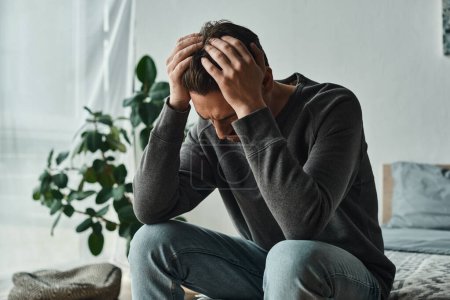 Aufgebrachte bärtige Mann in Casual Home tragen Kopf berühren, während auf dem Bett zu Hause sitzen, Depressionen