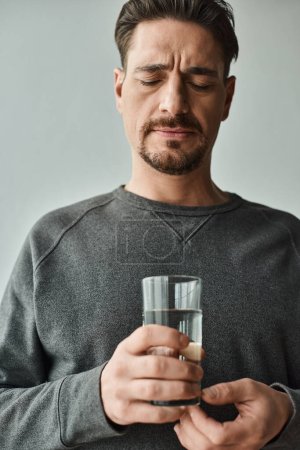 bärtiger Mann grimmig, während er unter Kopfschmerzen leidet und zu Hause ein Glas Wasser hält, unwohl
