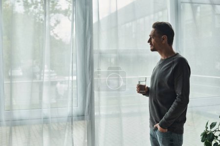 hombre barbudo reflexivo de pie con la mano en el bolsillo, mirando a la ventana y sosteniendo el vaso de agua