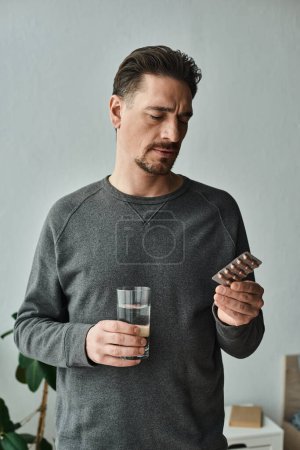 bärtiger Mann im lässigen grauen Pullover, der beim Betrachten von Medikamenten die Stirn runzelt und ein Glas Wasser in der Hand hält