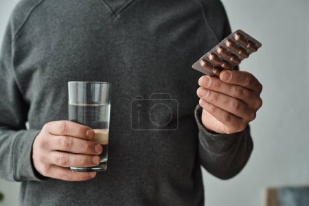 cropped photo of man in casual grey jumper holding glass of water and blister pack with medication