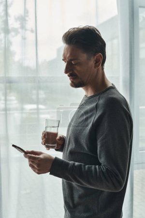 bärtiger Mann in lässigem grauen Pullover runzelt die Stirn und hält ein Glas Wasser in der Hand, während er Blisterpackung betrachtet