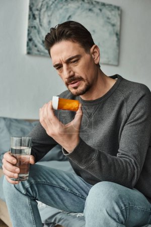 bearded man in grey jumper frowning and holding glass of water while looking at bottle with pills