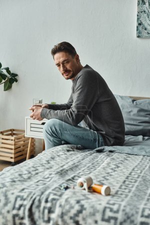 bearded man in casual attire holding glass of water and looking at medication on bed, remedy