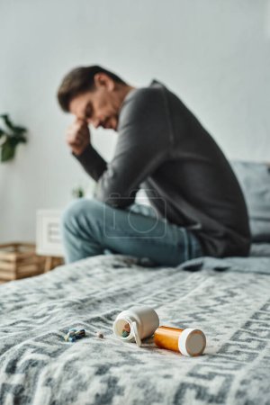 blurred and bearded man in casual attire sitting on bed with different medication, remedy