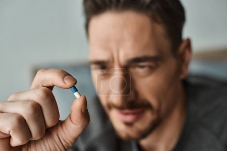 close up shot of bearded man in casual attire holding blue and white capsule in hand, medication