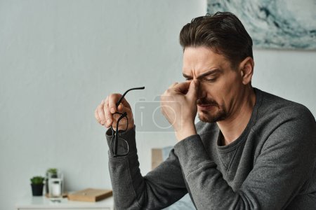 Foto de Hombre barbudo estresado en ropa casual sosteniendo gafas y tocando los ojos después del trabajo en casa, burnout - Imagen libre de derechos