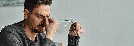 hombre barbudo estresado en ropa casual sosteniendo gafas y tocando los ojos después del trabajo en casa, pancarta