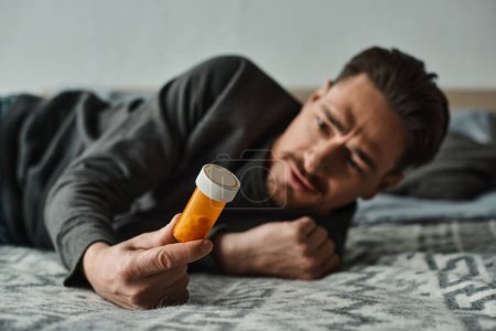 bärtiger Mann mit Bauchschmerzen und Liegen auf dem Bett in der Nähe der Flasche mit Medikamenten, Beschwerden