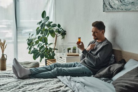 pensive man examining bottle with medication while lying on bed in bedroom, treatment plan