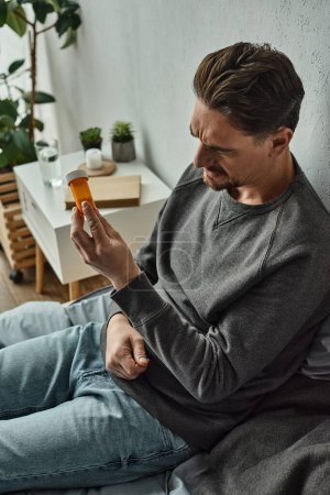 nachdenklicher Mann untersucht Flasche mit Medikamenten, während er im Schlafzimmer auf dem Bett sitzt, Behandlungsplan