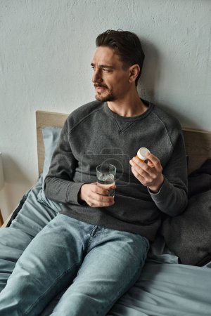 hombre reflexivo en suéter sosteniendo vaso de agua y botella con pastillas en el dormitorio, tratamiento