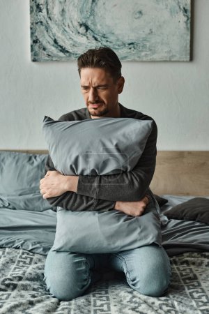 bärtiger Mann fühlt sich unwohl und hält Kissen, während er zu Hause auf dem Bett sitzt, beunruhigter Patient