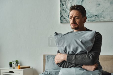 Photo for Bearded man feeling discomfort and holding pillow while sitting on bed at home, sad patient - Royalty Free Image
