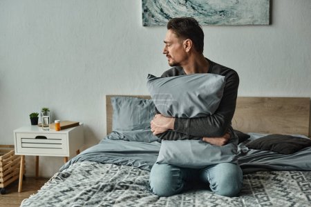 bearded man feeling discomfort and holding pillow while sitting on bed at home, modern interior