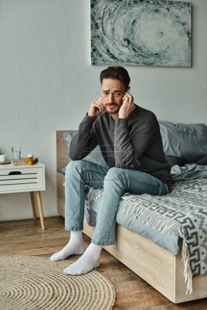 Besorgter bärtiger Mann mit Kopfschmerzen spricht auf Smartphone im modernen Schlafzimmer, Online-Beratung