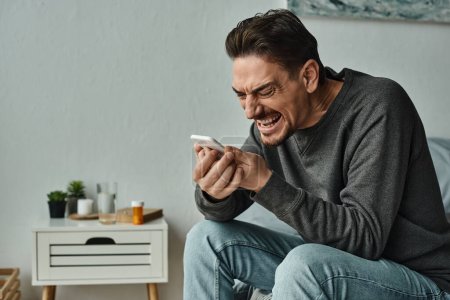 hombre barbudo en jersey gris gritando a su teléfono inteligente mientras está sentado en la cama en casa, emocional