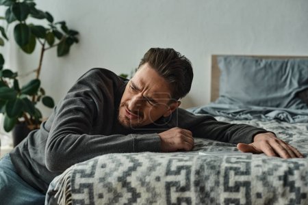 bärtiger Mann leidet unter Schmerzen und lehnt auf grauer Decke auf dem Bett im modernen Schlafzimmer, Stress