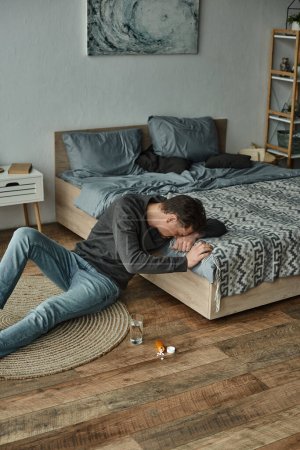 hombre barbudo estresado sentado en el suelo cerca de un vaso de agua y pastillas en el dormitorio, medicamentos